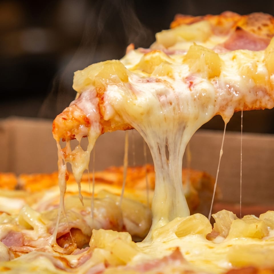 Close-up of a steaming Hawaiian pizza slice lifted above the pie, showing melted cheese, ham, pineapple, and browned crust.